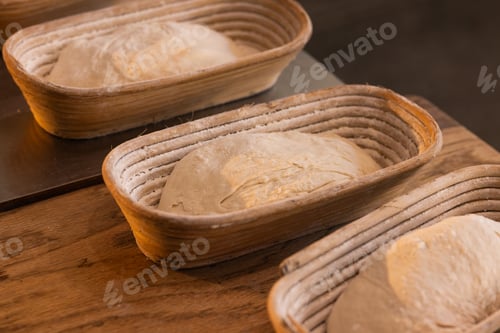 Preview: Oval woven bannetons holding three flour-dusted loaves of proofing dough on wooden countertop