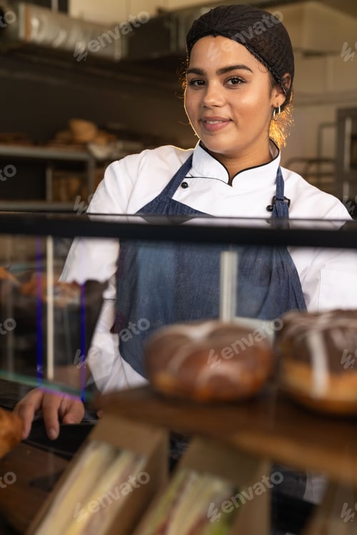 Preview: Adult African American woman baker in white jacket and blue apron standing behind glass pastry case
