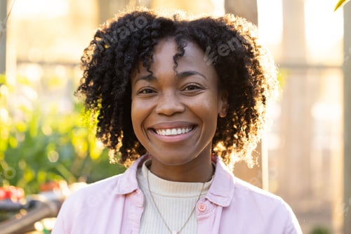 Preview: Smiling mid adult African woman standing in sunlit greenhouse wearing pink shirt and pendant