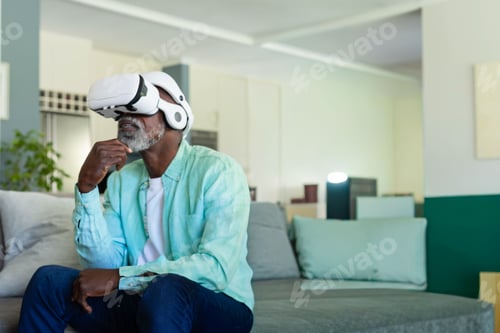 Preview: Mature African American man sitting on sofa at home, wearing white VR headset, shirt, resting chin