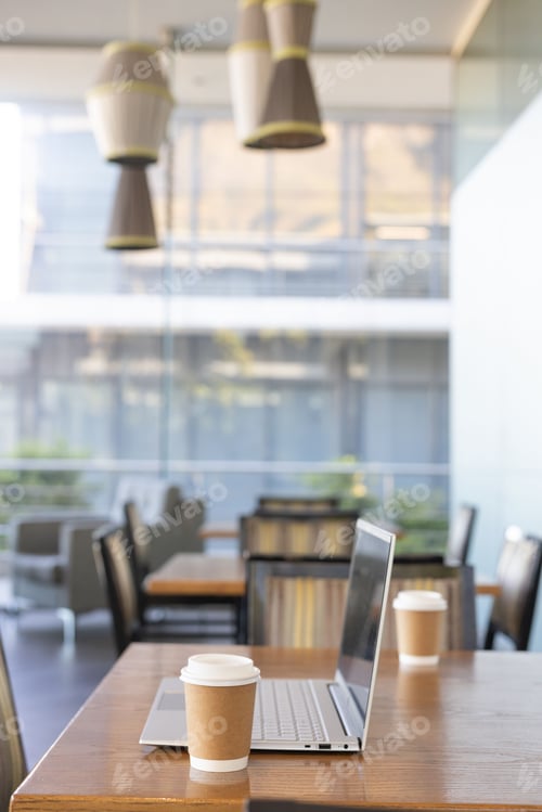 Preview: Open silver laptop sitting on wooden table with paper coffee cup near glass wall