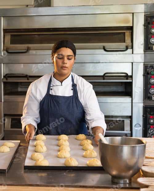 Preview: African American woman baker wearing chef coat holding tray with rolls by ovens and mixing bowl