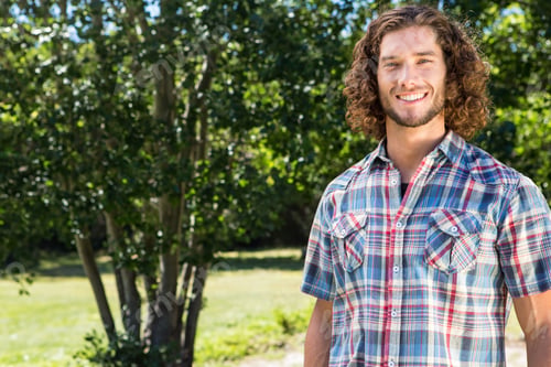 Preview: Male wearing plaid shirt standing smiling, facing camera on sunlit park lawn with tree, copy space