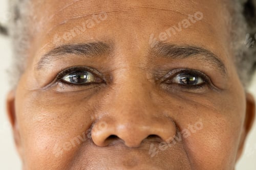 Preview: Senior woman's face is filling frame, showing textured skin and reflective eyes near pale wall