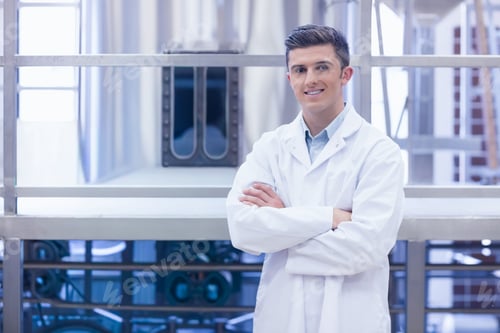 Preview: Male in 20s standing wearing white lab coat crossing arms facing camera in industrial plant