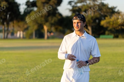 Preview: Adult male standing right on sports field holding clear water bottle, wearing white uniform