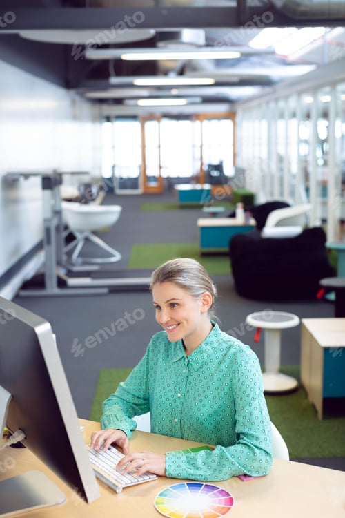 Preview: Large monitor is showing design layout while white keyboard is resting on wooden desk, open office