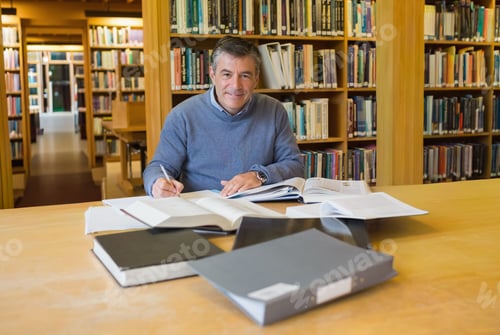 Preview: Mature man in sweater studying at wooden library table, reading textbooks and taking notes with pen