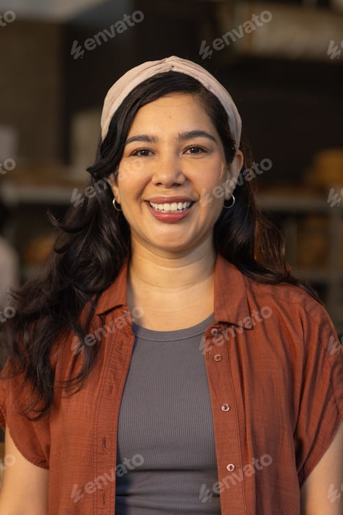 Preview: Woman smiling, looking at camera in storeroom shelving with rust shirt, headband and hoop earrings