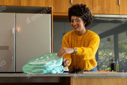 Preview: Adult woman smiling in mustard sweater organizing teal backpack at kitchen island by cutting board