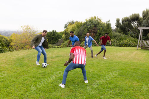 Preview: Five male friends playing soccer on grassy lawn, man in striped jersey defending near soccer ball