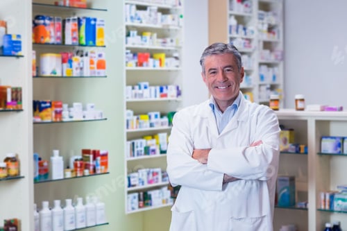 Preview: Mature male pharmacist standing with arms crossed wearing white lab coat at pharmacy shelves