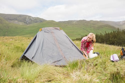 Preview: Adult woman kneeling beside gray dome tent on grassy moor, securing guyline with stake, copy space
