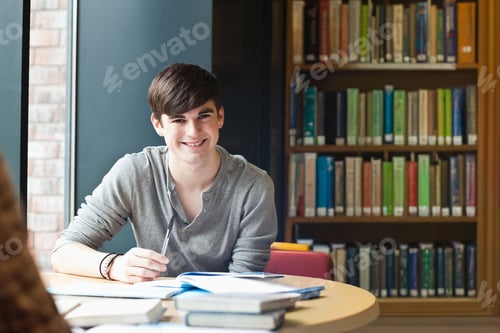 Preview: Teenage male student sitting at round study table near tall window holding pen over open textbook