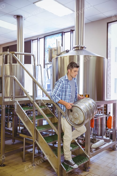 Preview: Man carrying stainless keg down metal stairs inside brewery by fermenters, wearing plaid shirt