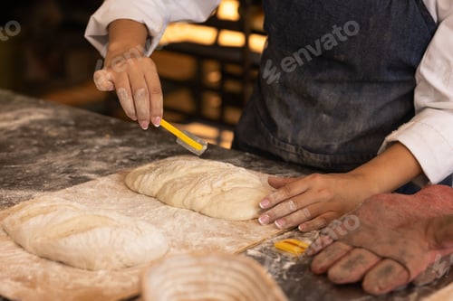 Preview: Adult African American woman in white chef jacket scoring loaves on floured board with yellow blade