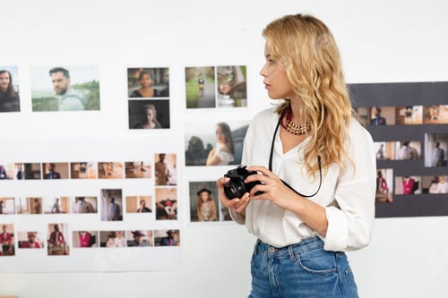 Preview: Adult blonde woman in white blouse holding camera inspecting prints on white wall, copy space