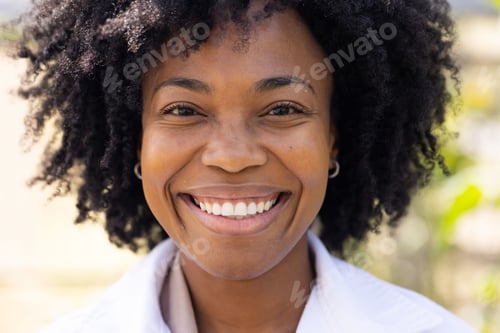 Preview: Mid adult African woman smiling, facing camera outdoors wearing silver hoop earrings, collared top