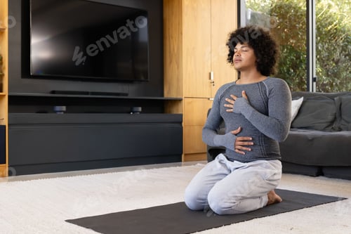 Preview: African American man kneeling, breathing on yoga mat at home, wearing gray athletic top, copy space