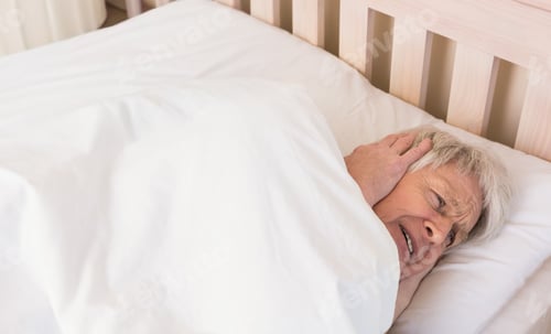 Preview: Senior man lying sideways on bed clutching left ear under white pillow and duvet, copy space
