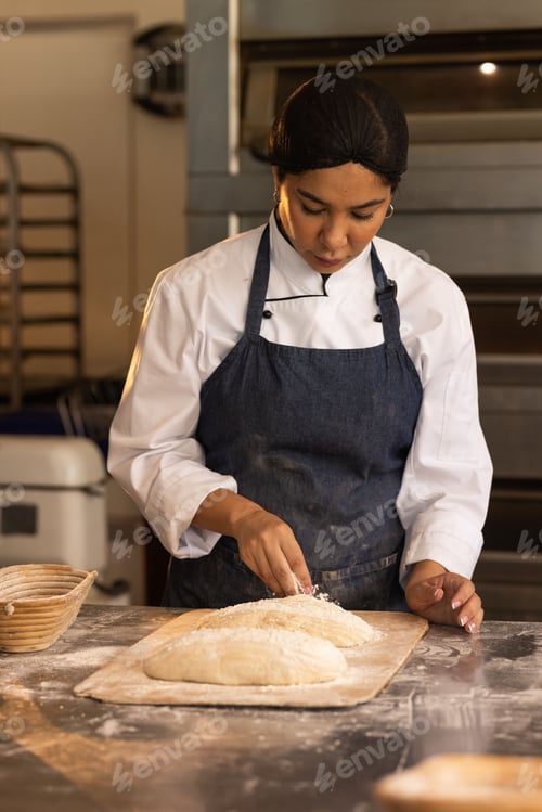 Preview: Female baker wearing chef coat and apron shaping two oval loaves on peel in bakery kitchen