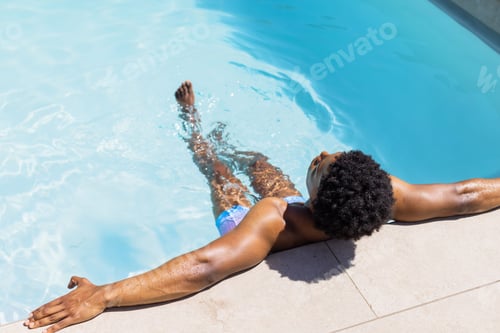 Preview: African American man reclining on stone coping at pool in blue trunks causing ripples, copy space