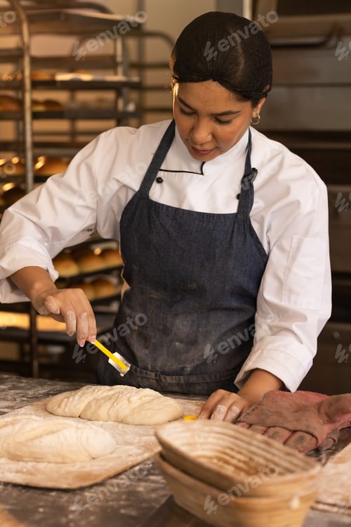 Preview: Female baker in chef whites, cap brushing glaze onto loaves using yellow brush on floured bench