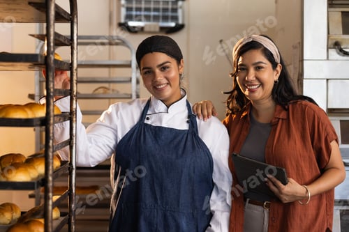 Preview: Female bakery coworkers with baker wearing coat and apron, manager holding tablet by rack of rolls