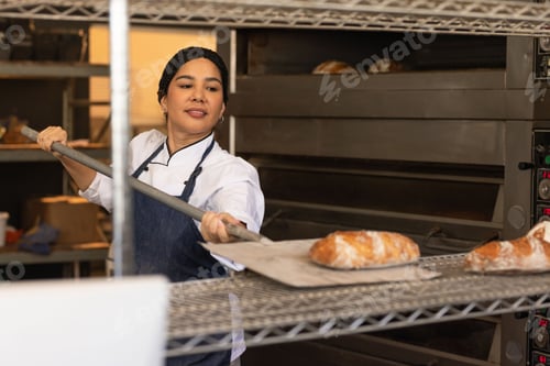 Preview: Female baker wearing chef jacket dark apron moving crusty loaves with metal peel in bakery