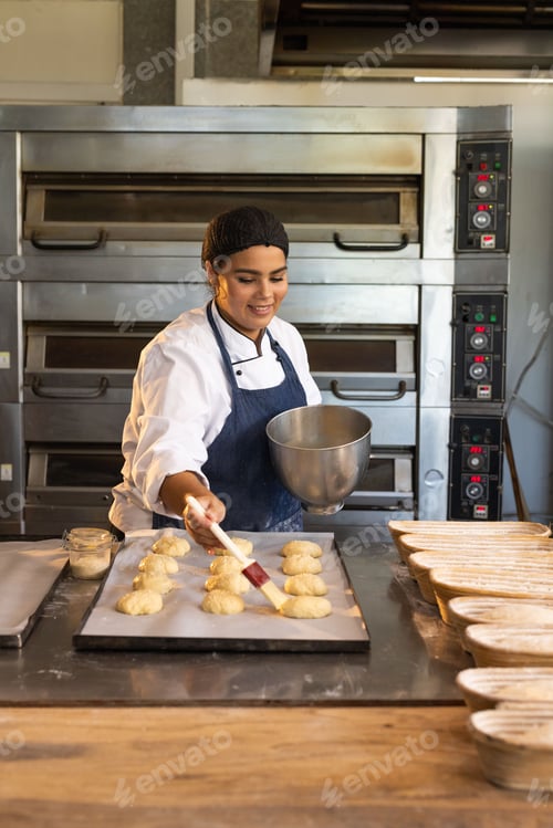 Preview: African female baker in chef-jacket brushing glaze onto dough on-parchment at steel bench with-bowl