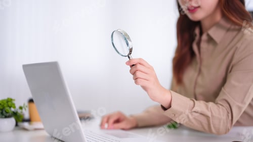 Preview: Asian businesswoman holding magnifying glass analyzing research data using laptop computer working i