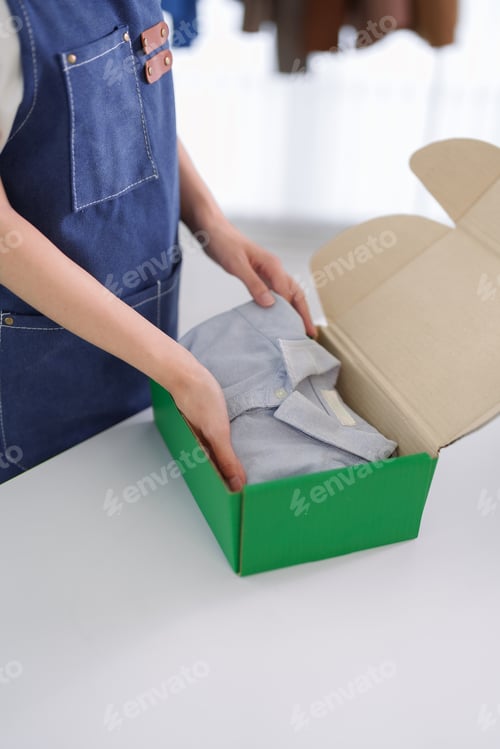 Preview: Asian women hands packing folded clothing into green box preparing shipment for online store ecommer