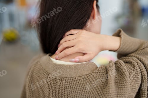 Preview: Adult woman touching neck and shoulder experiencing muscle tension pain while wearing beige knit swe