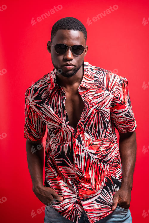 Preview: Cool African American man in tropical shirt and sunglasses looking at camera on red background