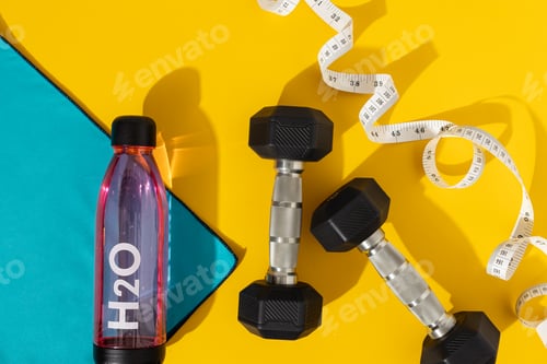 Preview: Flat Lay of Water Bottle and Blue Towel Next to Dumbbells With Measuring Tape on Yellow Background.