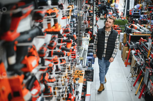 Preview: Portrait of young bearded man buying modern electric drill while standing in tool. Hardware construc