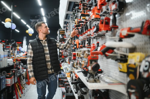 Preview: Portrait of young bearded man buying modern electric drill while standing in tool. Hardware construc