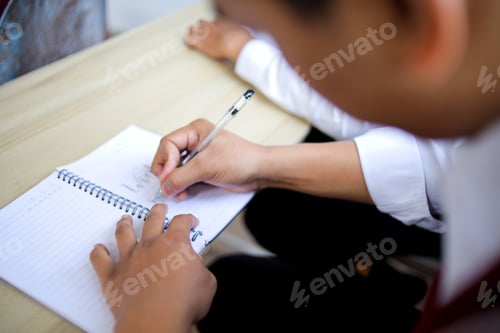 Preview: Over Shoulder Closeup of Student Writing with Clear Gel Pen in Spiral Notebook on Wooden Desk