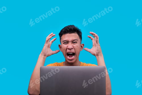 Preview: Angry Asian Man Screaming Behind Laptop with Hands Raised, Expressing Frustration or Stress
