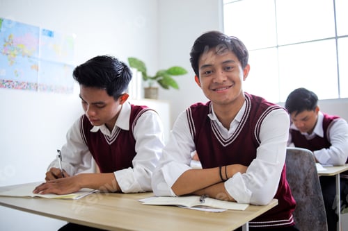 Preview: Asian High School Student Smiles at Camera With Arms Crossed While Classmates Study