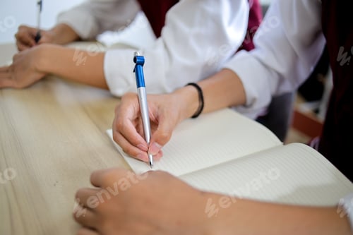 Preview: Closeup Shot of Students Hand Holding Blue Pen and Writing in Notebook on Wooden Desk During an Exam