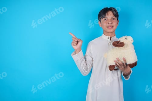 Preview: Smiling Asian Muslim Boy Holding Sheep Doll And Pointing Up At Copy Space On Blue Background