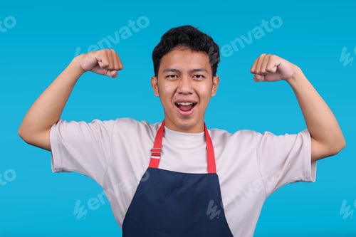 Preview: Enthusiastic Young Asian Man in Blue Apron Flexing Both Arms