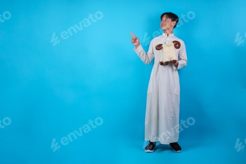 Preview: Full Body Shot Of Smiling Asian Muslim Boy Holding Sheep Doll and Pointing Up At Copy Space