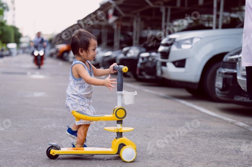 Preview: Cute 2-year-old Asian boy pushing his yellow kick scooter near a parking lot, raising awareness abou