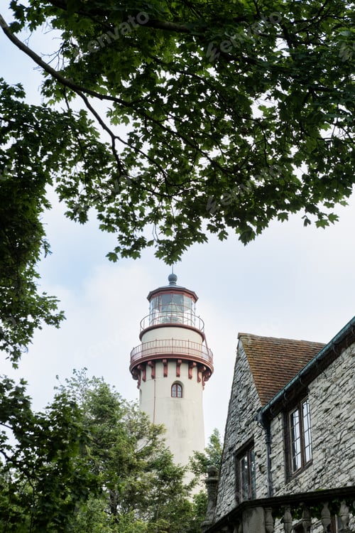 Preview: Lighthouse stands tall among trees near a stone building on a cloudy day by the coast