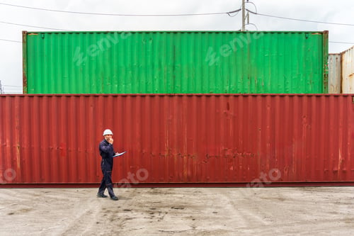 Preview: Male logistics cargo worker talking on walkie talkie while walking past stacked shipping containers