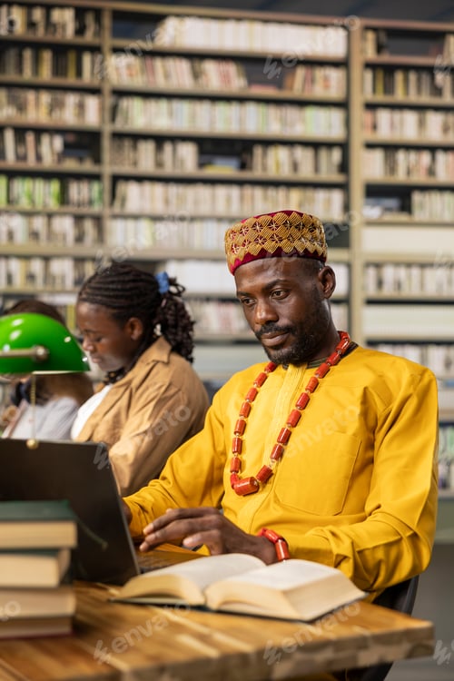 Preview: African man in traditional clothes working on laptop beside open book