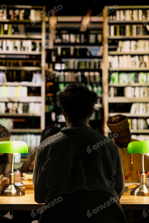 Preview: Back view of young adult student sitting at dim library table with peers