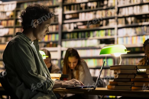 Preview: Student working on laptop at dim library table while classmates study nearby